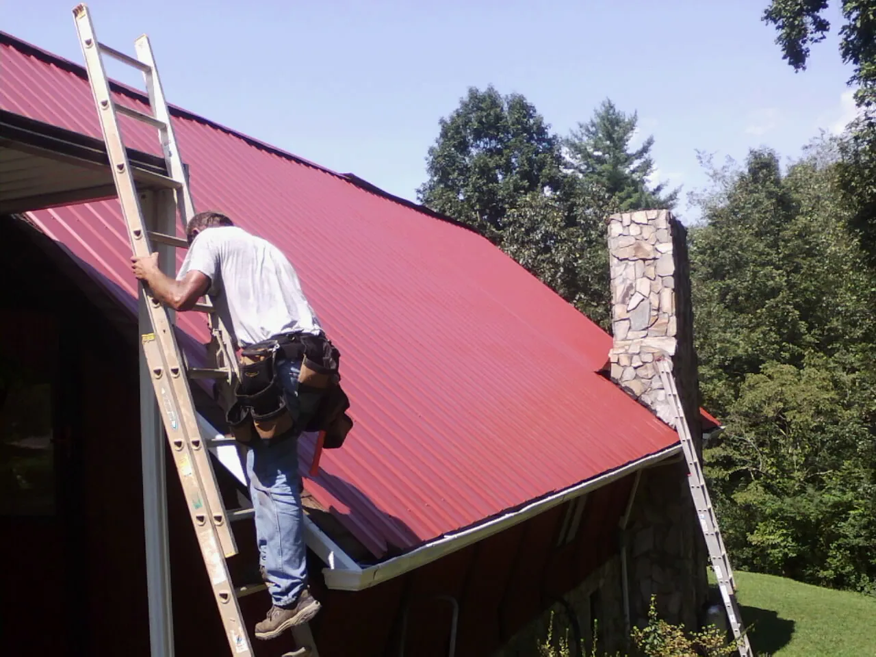 Roofer on a ladder beside a red metal roof and stone chimney — Soffit & Fascia Repair by Roofing Brothers in Gretna